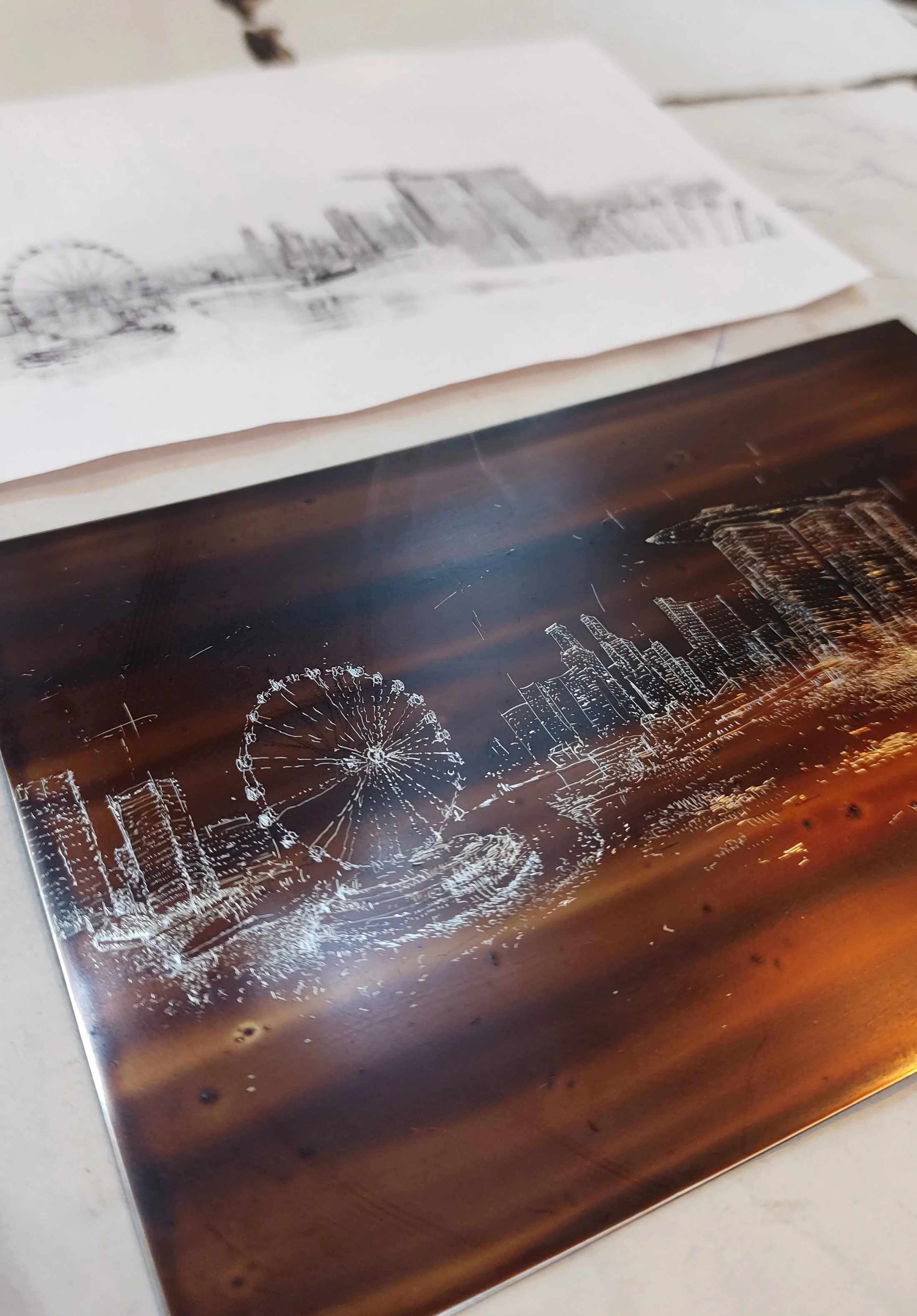 Engraving plate with the Singapore skyline and sun, beside a paper sketch